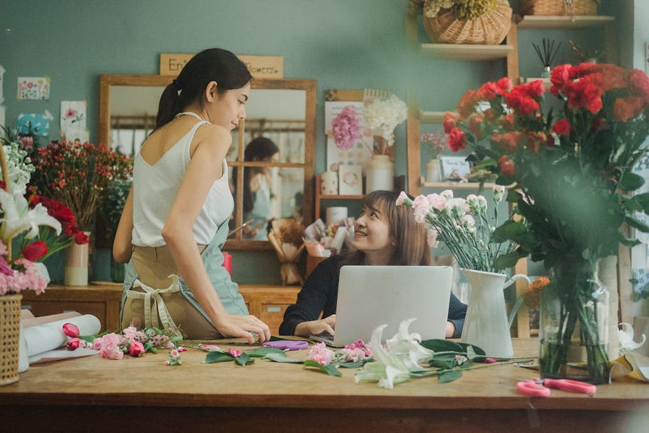 A young woman with long brown hair, wearing a black top, sits at a wooden table in a floral shop, smiling up at a female staff member with dark hair tied back, dressed in a sleeveless white top and a 