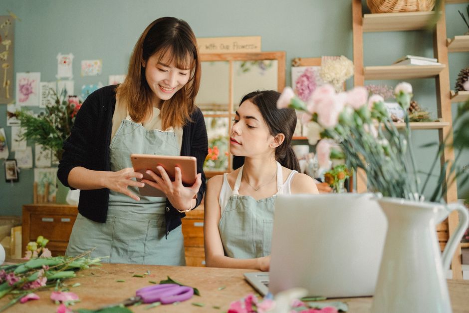 A floral shop scene featuring two women working with fresh flowers. The woman on the left, with shoulder-length brown hair and a black cardigan, is smiling and holding a tablet, possibly reviewing a f