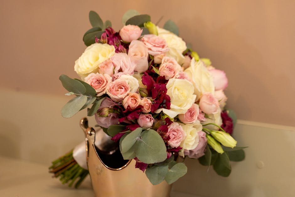 A bouquet of fresh flowers arranged in a rounded style, featuring light pink roses, creamy white roses, and deep burgundy flowers, complemented by green eucalyptus leaves. The bouquet is wrapped in wh