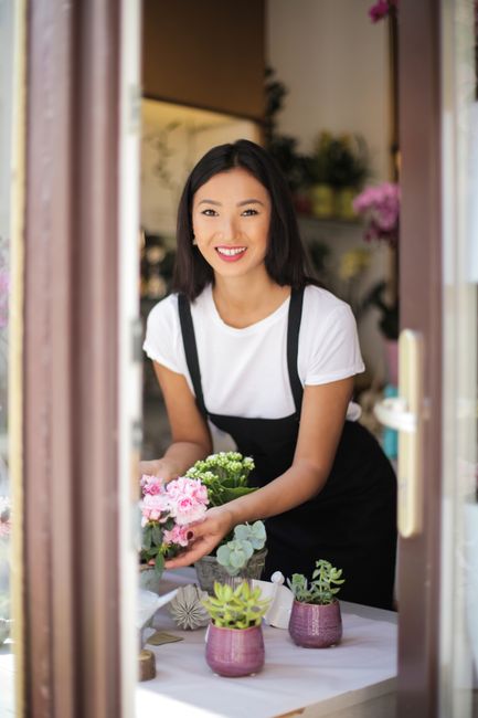 A young woman with shoulder-length black hair and a bright smile, dressed in a white t-shirt and black apron, is standing behind a table inside a floral shop. She is holding a bouquet of fresh pink ca