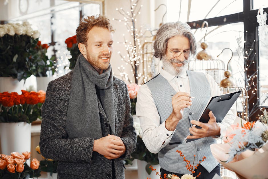 A professional florist with gray hair and glasses, wearing a light gray vest and white shirt, is using a tablet to manage flower arrangements inside a bright flower shop. Next to him, a young man with