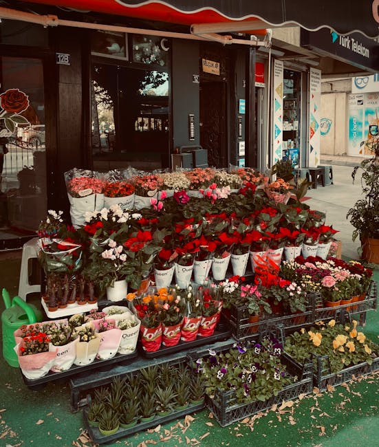 An outdoor display of fresh flowers in front of a flower shop, featuring a variety of floral arrangements with red roses, pink tulips, white daisies, and mixed seasonal blooms. The flowers are arrange