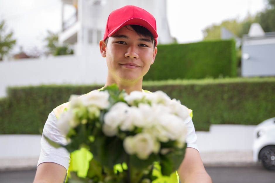 A delivery person wearing a red cap and a high-visibility vest is smiling and holding a bouquet of fresh white roses and green foliage, with a blurred background of residential houses and a car, indic