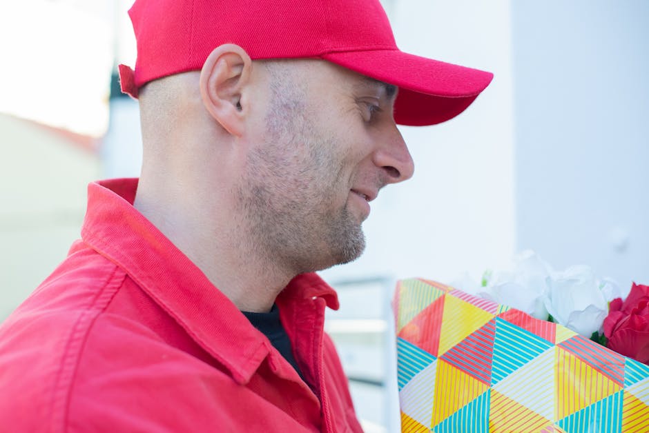 A delivery person dressed in a red cap and matching red jacket holds a vibrant floral gift wrapped in colorful, geometric-patterned paper, with white tissue paper and pink flowers visible inside. The 